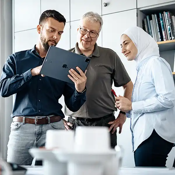 Drei Mitarbeitende – ein junger Mann, ein älterer Herr und eine Frau mit Kopftuch – schauen gemeinsam auf ein Tablet in einem modernen Büro
