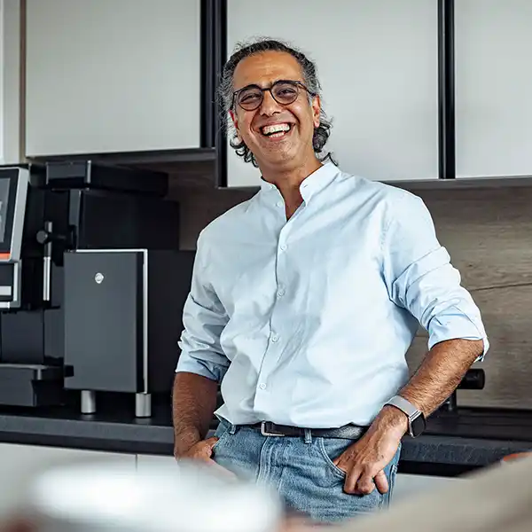 Ein lächelnder Mann mittleren Alters mit Brille, weißem Hemd und Jeans lehnt entspannt an einer Büroküche mit Kaffeemaschine im Hintergrund.